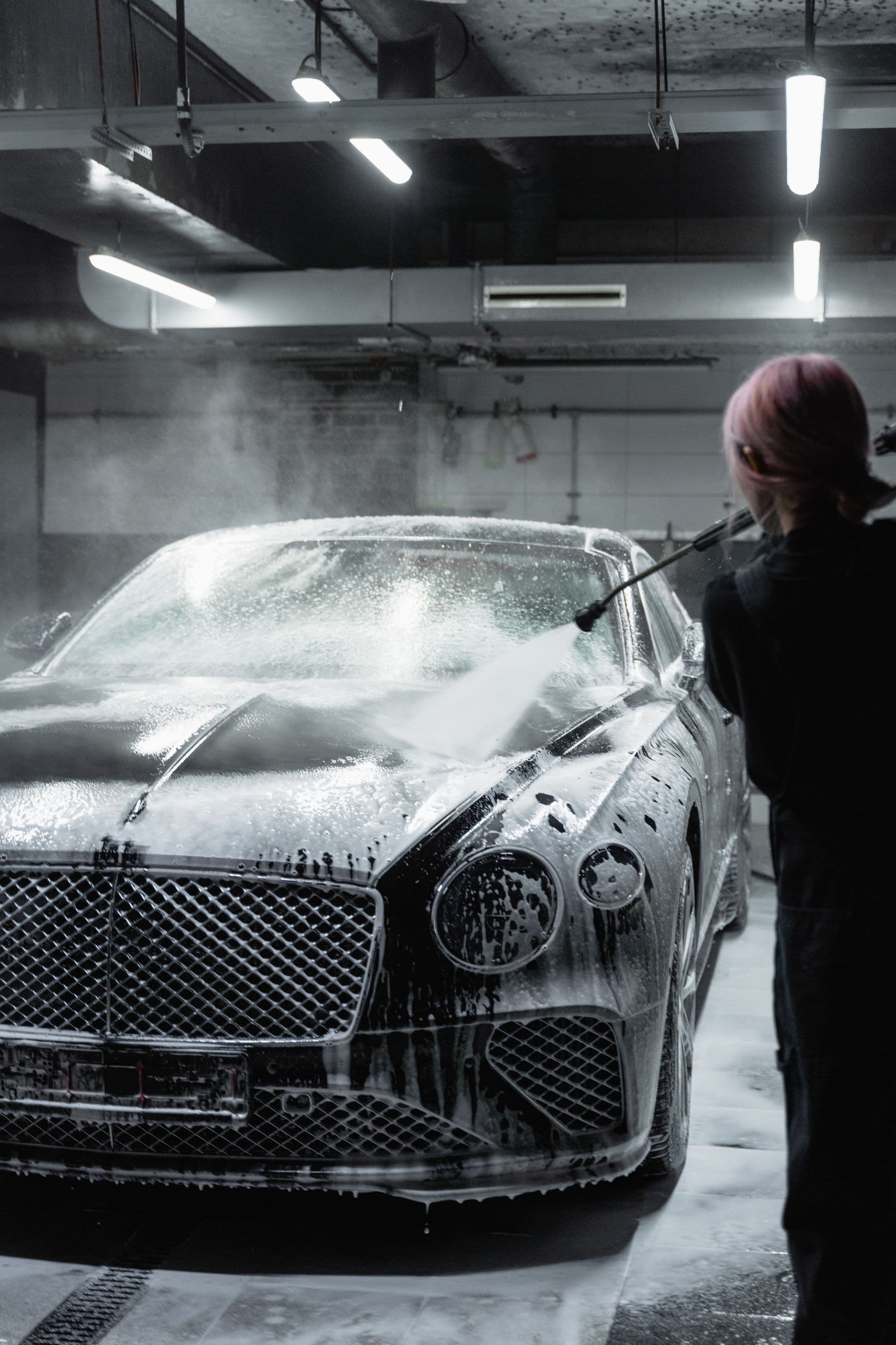 Technician rinsing thick foam from a dark luxury coupe in a covered parking bay